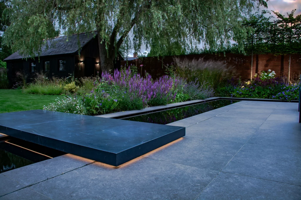 Sleek stone bridge lit from beneath beside evening planting.