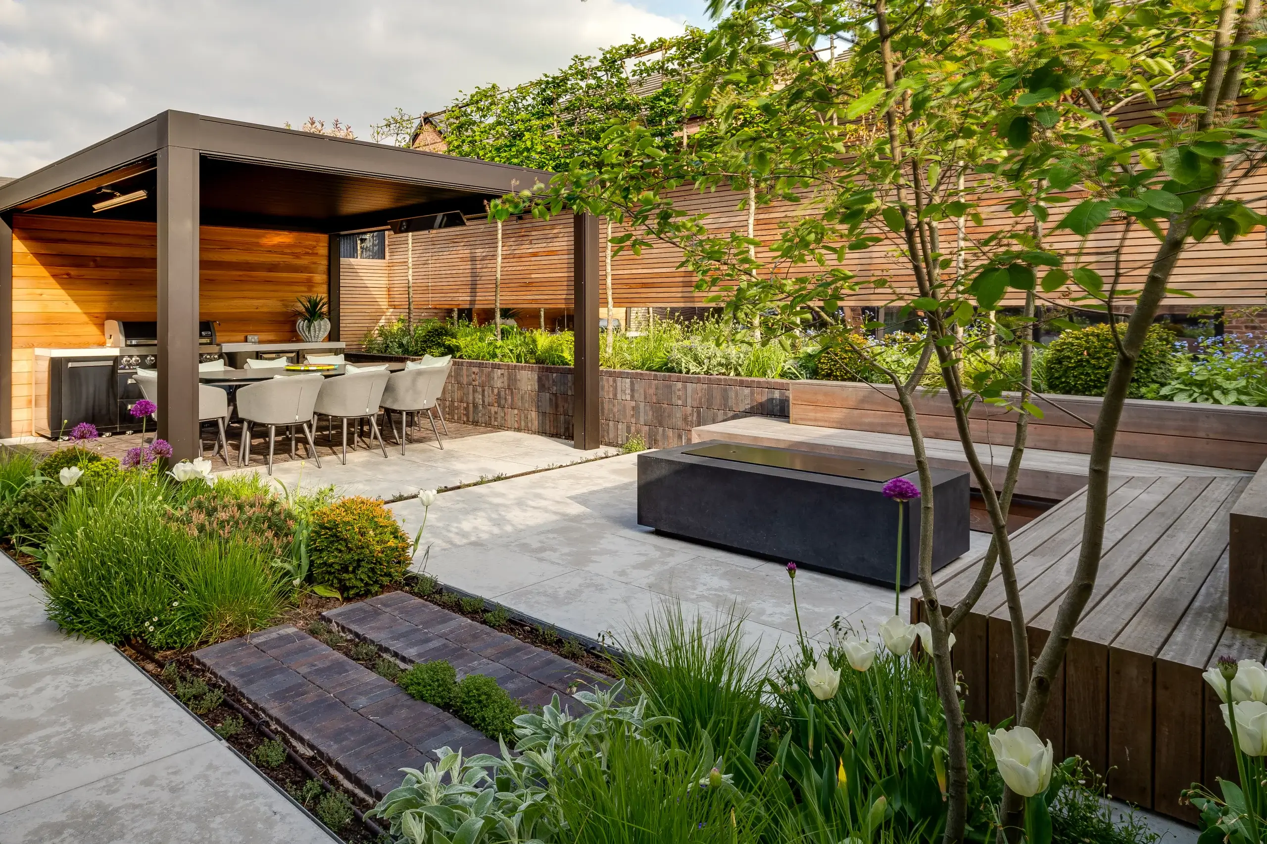 A modern landscaped garden by Base Squared featuring a covered outdoor dining area with a built-in barbecue, a sleek firepit with wooden bench seating, and lush planting with tulips and alliums.