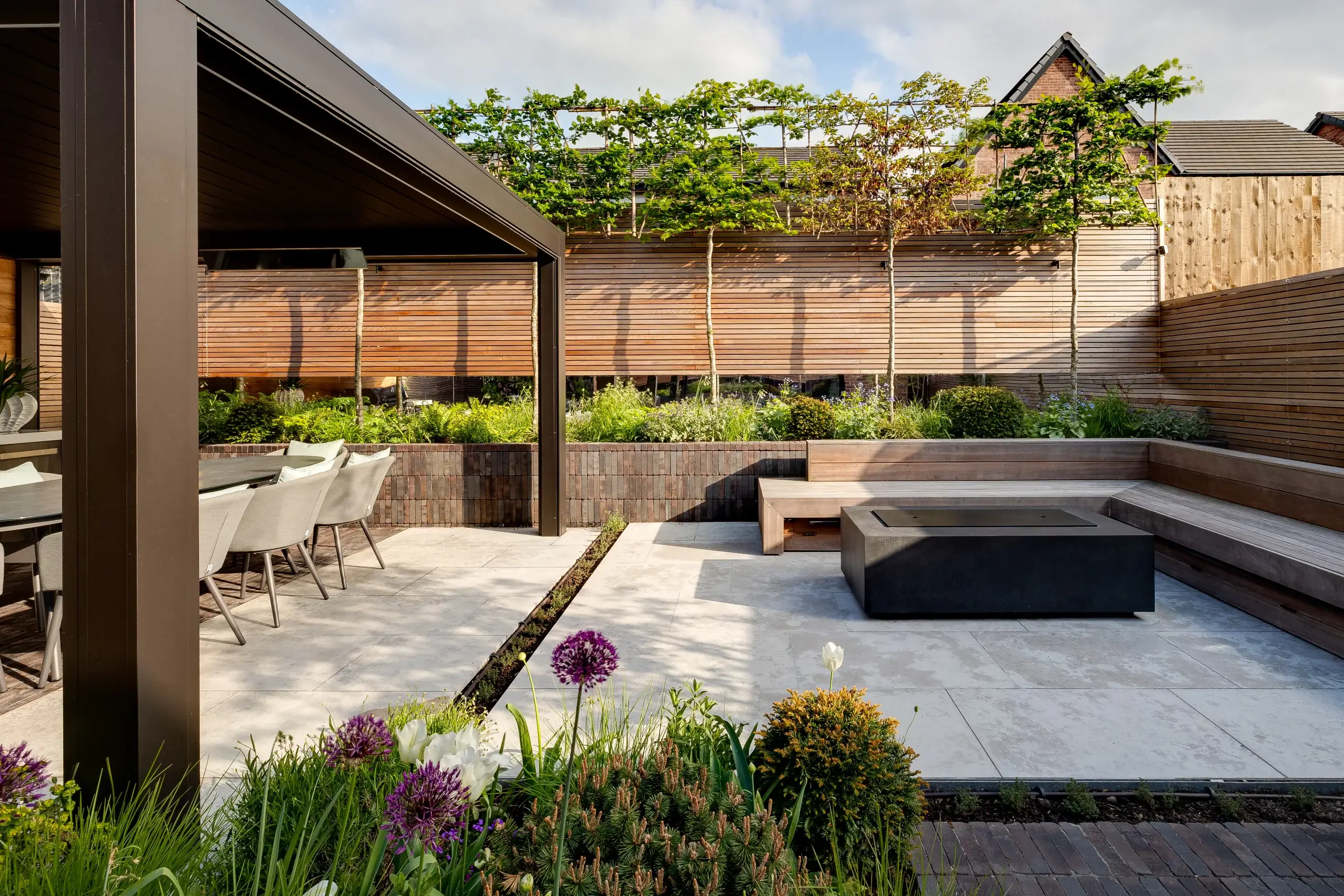 A landscaped modern garden by Base Squared featuring an outdoor dining area under a pergola, built-in wooden bench seating around a firepit, raised planting beds, and lush greenery with flowering alliums.