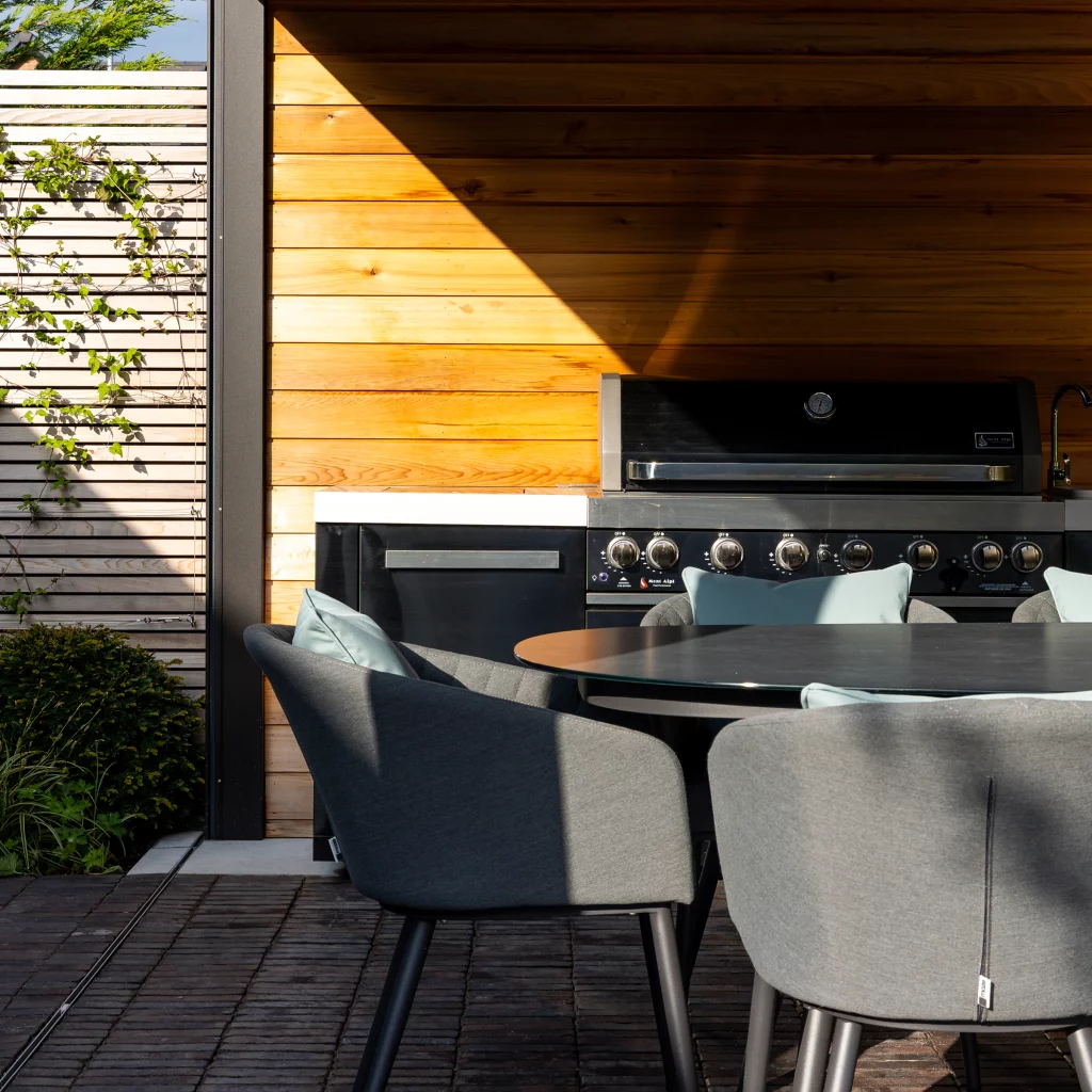 A modern outdoor kitchen with a sleek black gas barbecue, wooden cladding, and a round dining table surrounded by grey chairs, designed for stylish alfresco entertaining.