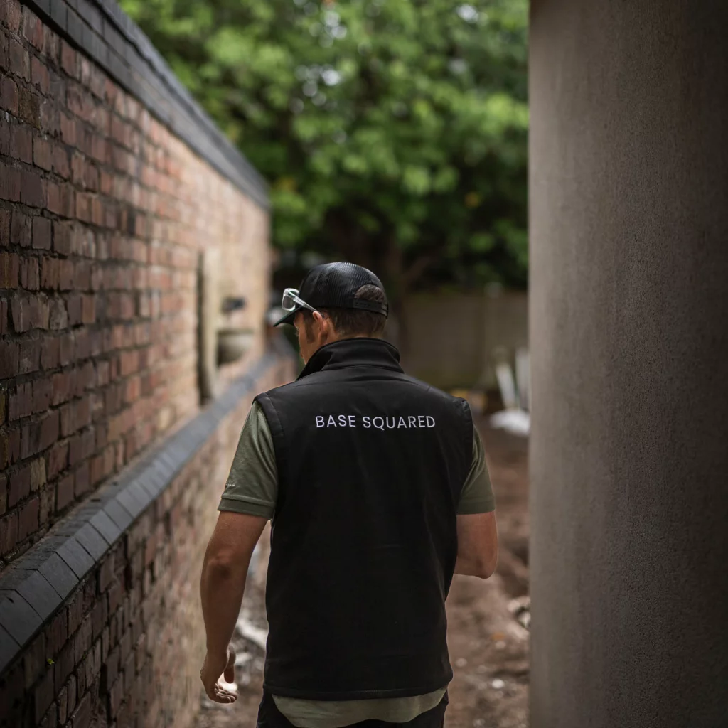 A Base Squared landscaper walks along a garden pathway in progress, wearing branded workwear with “Base Squared” on the back, next to a brick wall and greenery.