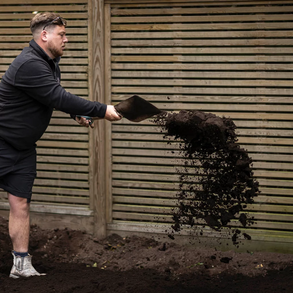 A Base Squared landscaper shovels rich soil across a garden bed, framed by a modern wooden slatted fence in the background.