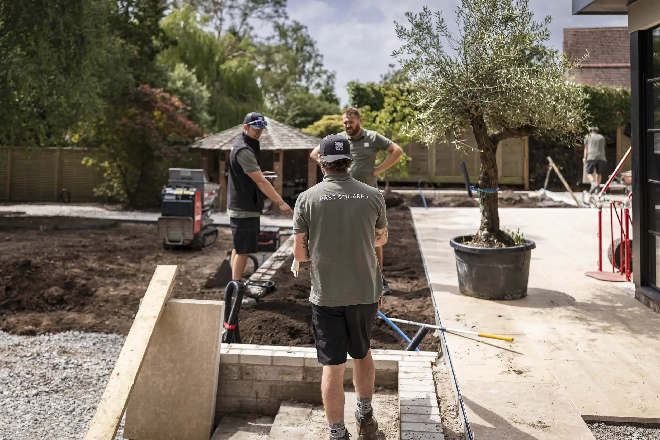 Base Squared landscaping team working on an outdoor project, with one team member in the foreground wearing a branded shirt and others discussing plans by a potted tree and construction equipment."