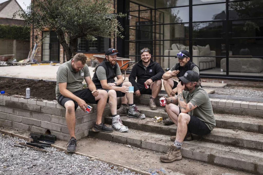 Base Squared landscaping team taking a break, sitting on steps outside a project site, sharing drinks and laughing together.