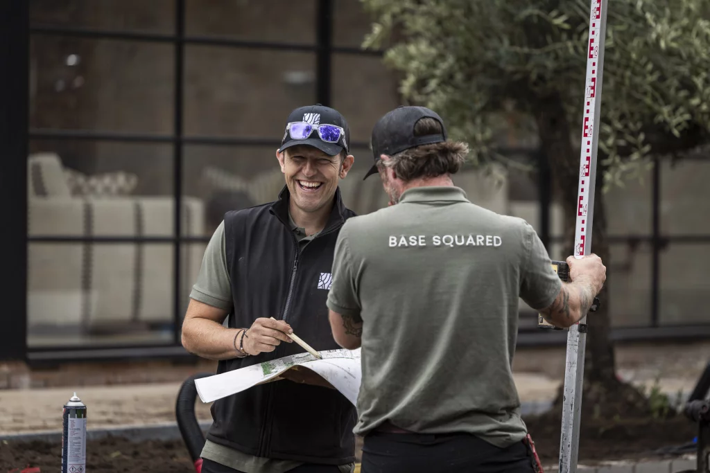 Two landscape designers from Base Squared working outdoors, smiling and reviewing plans together, with Tom holding drawings and the other holding a measuring staff.