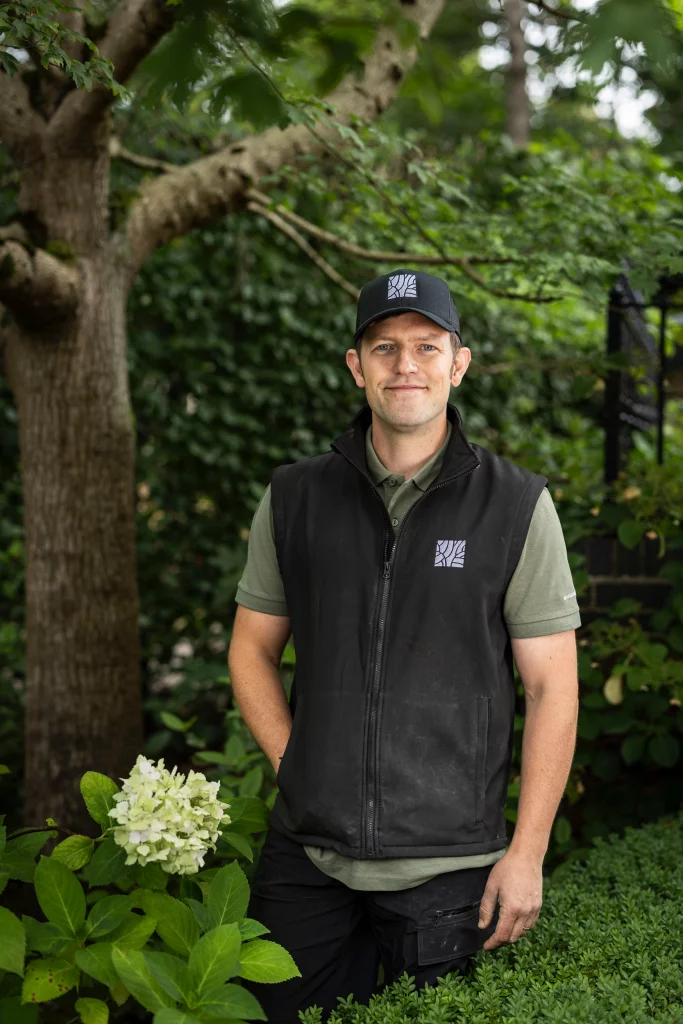Portrait of a landscaper wearing Base Squared uniform, standing among lush greenery with a hydrangea in the foreground.