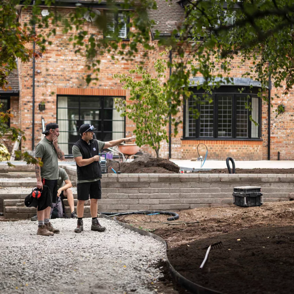 Two landscapers from Base Squared standing on a gravel pathway at a residential garden project, with one pointing towards the work area as they discuss progress. A brick house and newly planted tree are visible in the background.