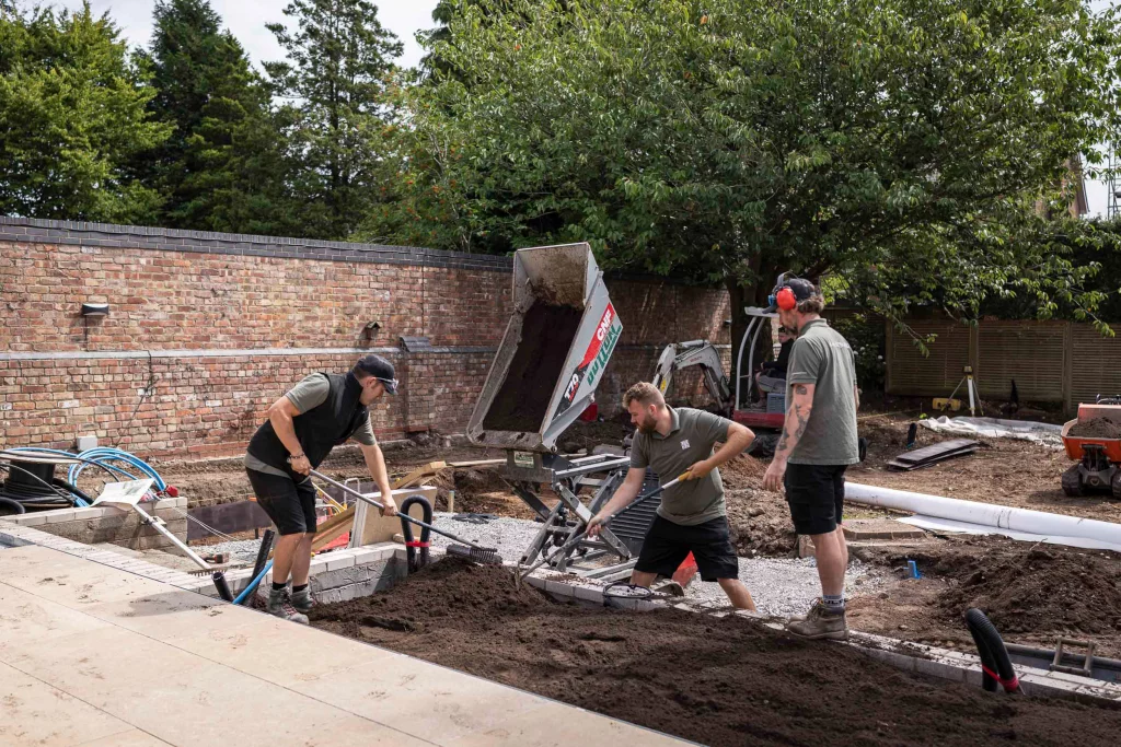 Three Base Squared landscapers level soil with rakes beside a brick wall, as a mini dumper unloads fresh topsoil into the garden under construction.