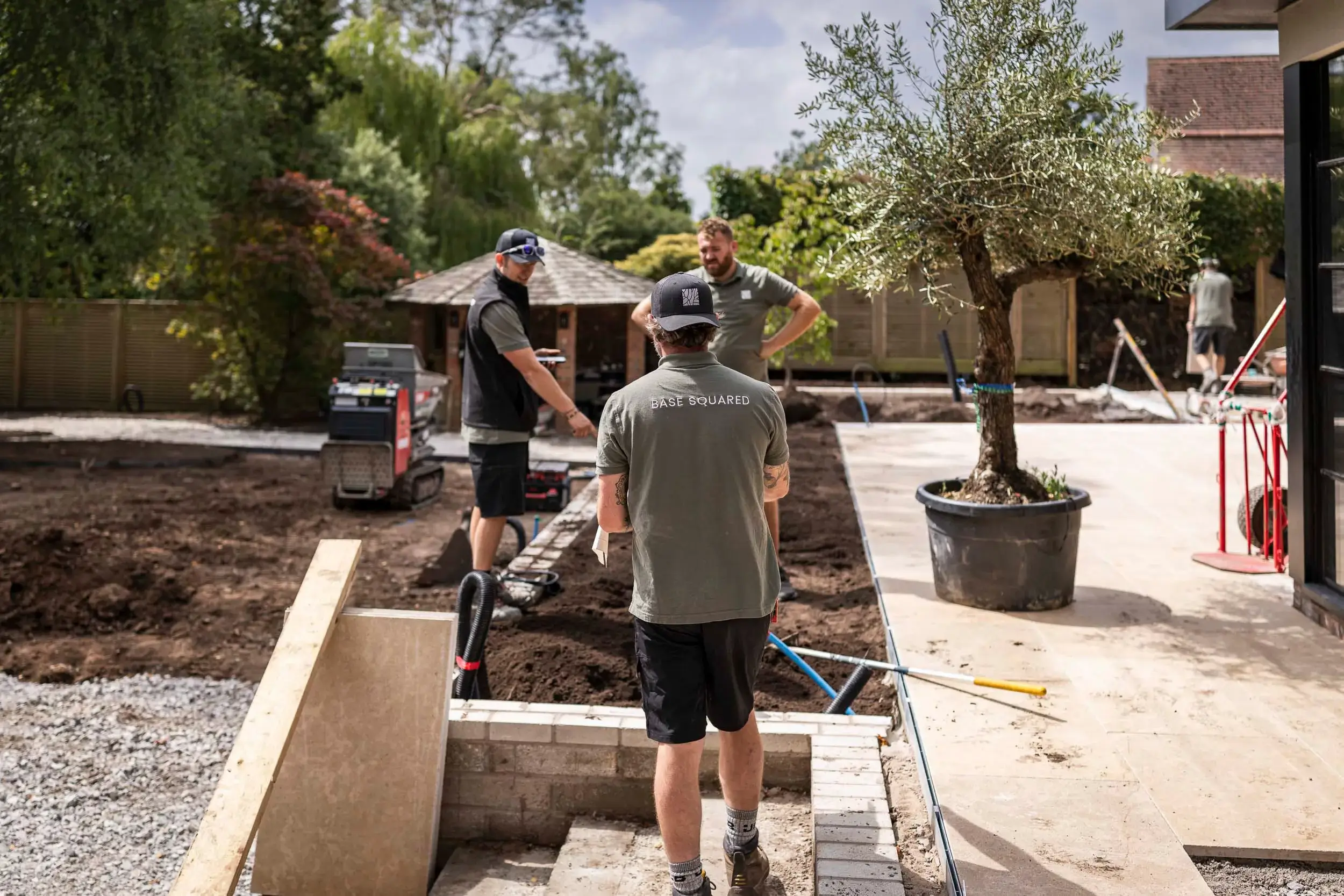 Base Squared landscaping team working on an outdoor project, with one team member in the foreground wearing a branded shirt and others discussing plans by a potted tree and construction equipment."