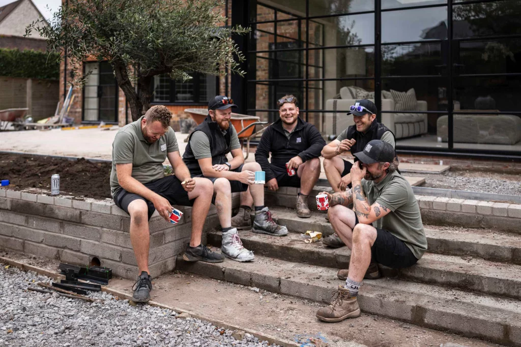 Base Squared landscaping team taking a break, sitting on steps outside a project site, sharing drinks and laughing together.