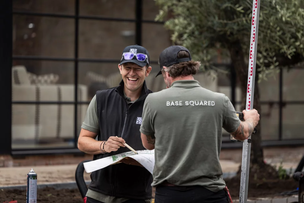 Two landscape designers from Base Squared working outdoors, smiling and reviewing plans together. With Tom holding drawings and the other team member holding a measuring staff.